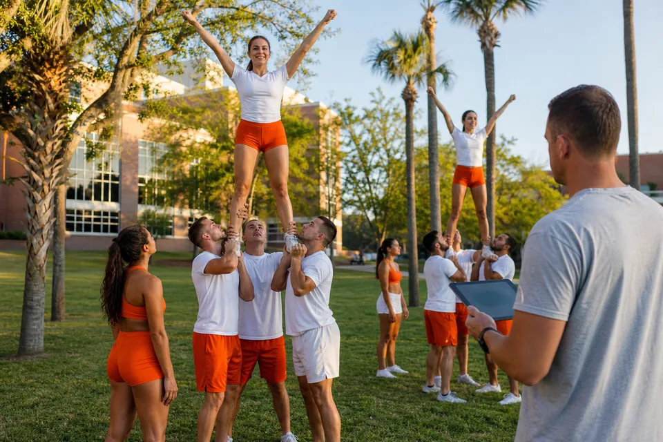 A college cheer squad practicing a partner stunt on a campus lawn lined with palm trees