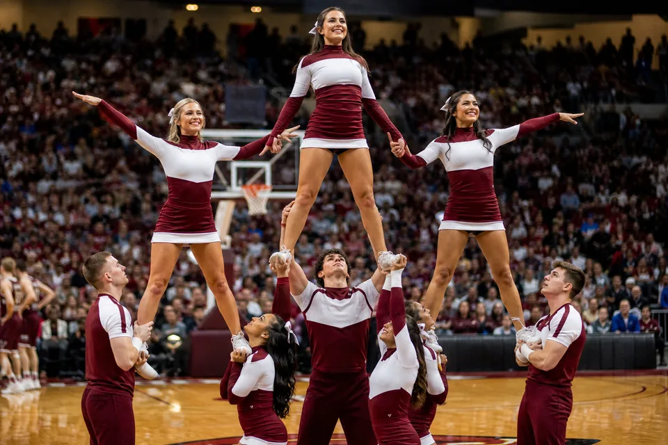 A coed college cheer squad in blue and white uniforms building a pyramid on a basketball court sideline